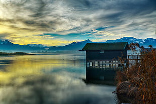 grey and green wooden house on center field water with mountain hills under cloudy sky during daytime