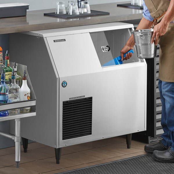 A man using a blue scoop to pour ice into a Hoshizaki undercounter ice machine.