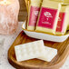 A white bar of Noble Eco Novo Natura soap on a wooden surface next to red and yellow packages of soap.