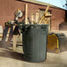 A man using a saw to cut wood into a grey Rubbermaid BRUTE trash can.