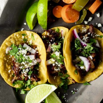 Overhead of pork carnitas served in corn tortillas with lemon wedges and pickled carrots and jalapeños on the side.