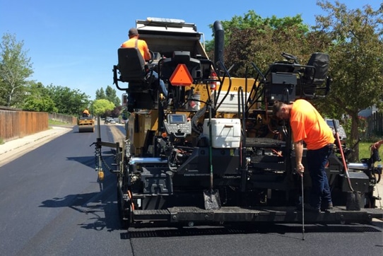 Workers in orange shirts operating a paving machine on a road under construction. A steamroller is visible in the background. The area is surrounded by trees and a wooden fence.