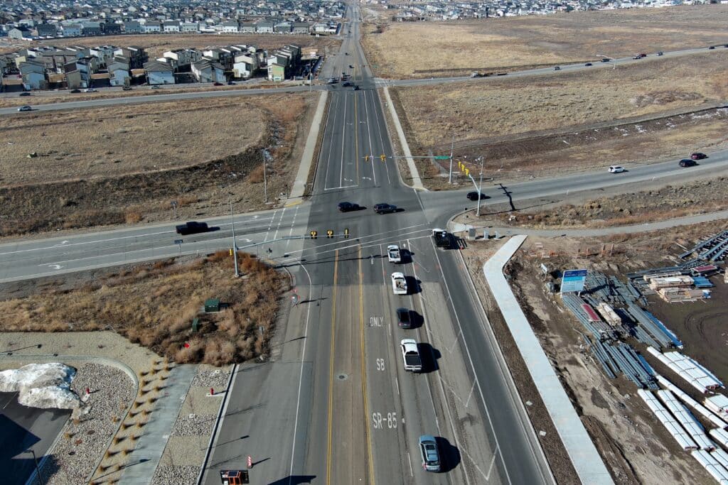 Aerial view of a wide intersection with multiple cars on the road. Surrounding the intersection are fields, a construction site, and residential houses in the distance. The landscape appears dry with minimal vegetation.