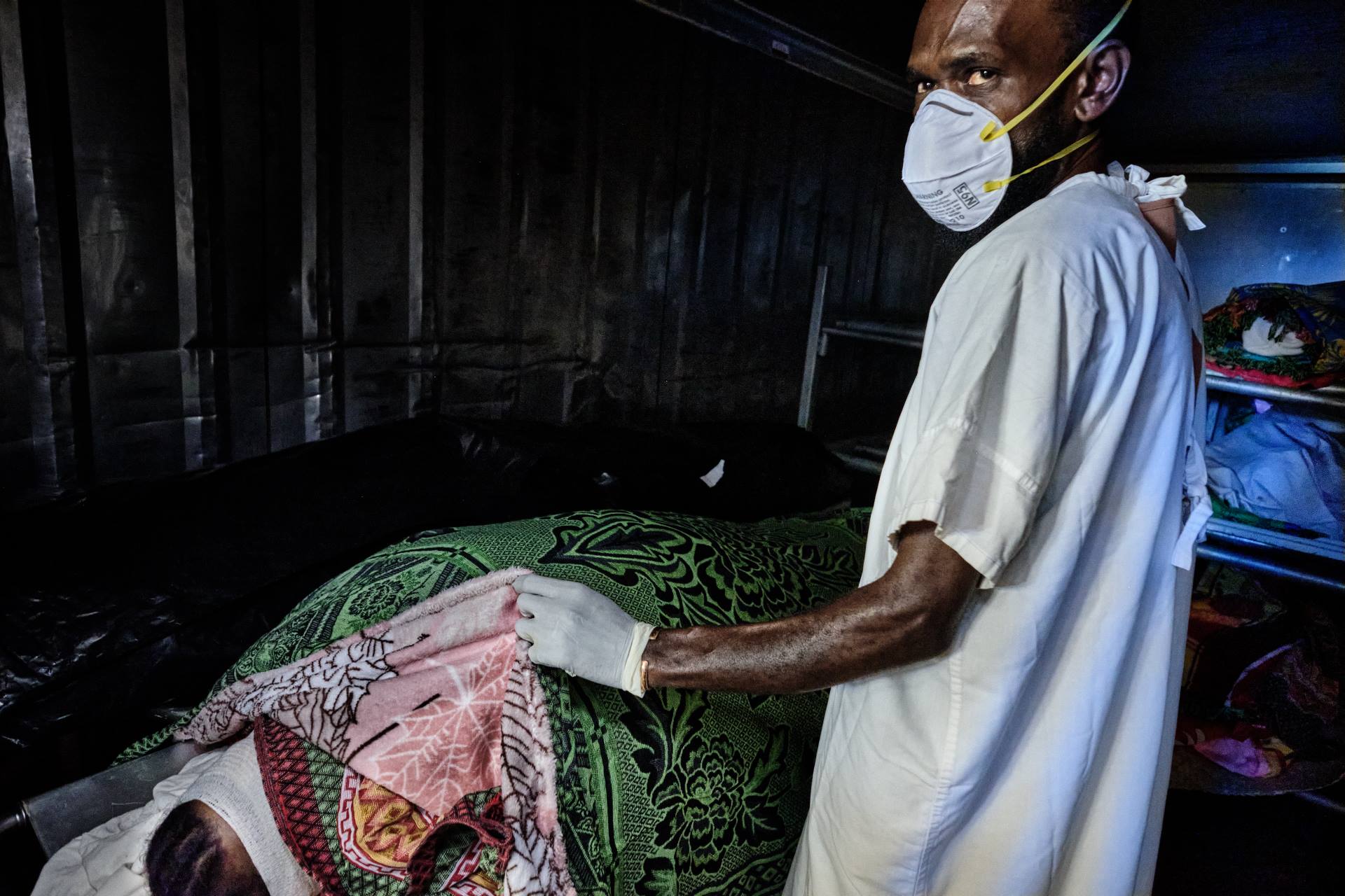 A man wearing a face mask cover a body in a morgue