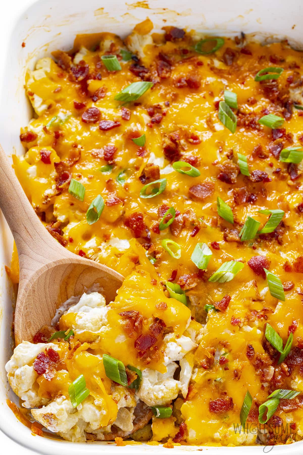 Scooping loaded cauliflower casserole with wooden serving spoon.