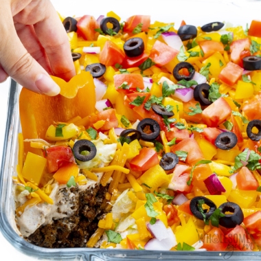 Taco dip recipe dunked with a hand holding a sliced bell pepper.