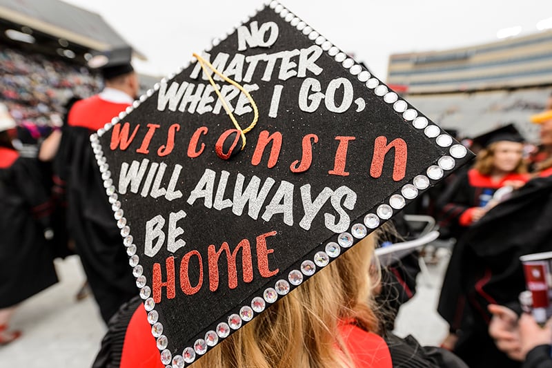 Graudate wearing a mortarboard with lettering on it reading 'No matter where I go, Wisconsin will always be home'