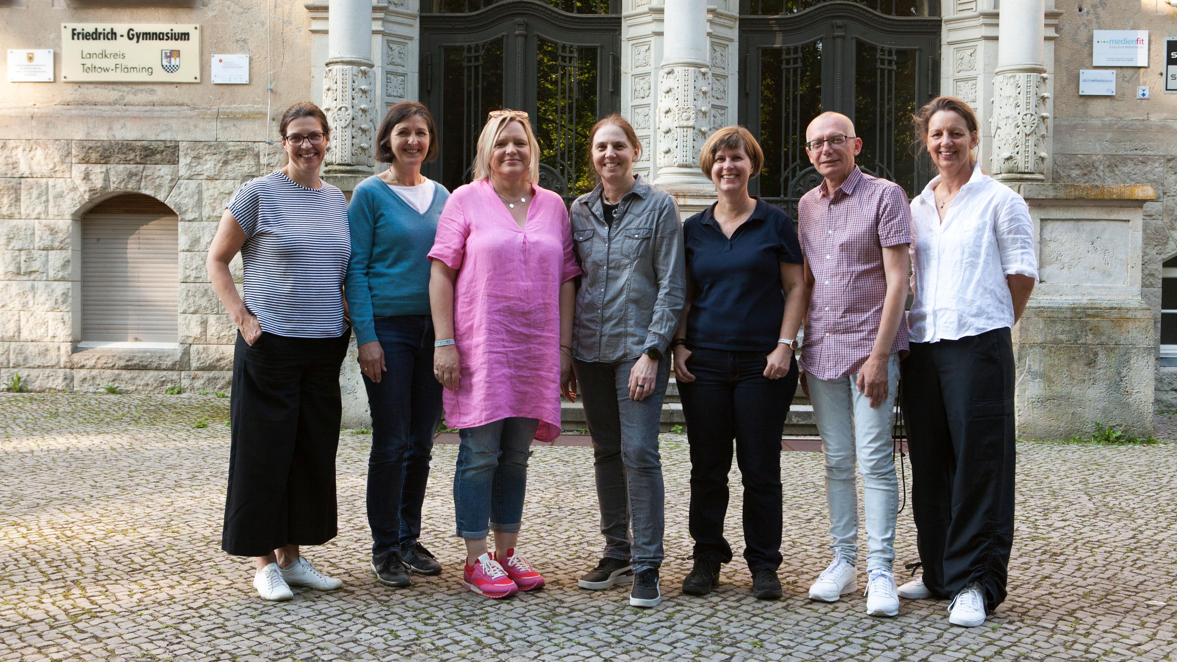 Sieben Erwachsene stehen vor einer Schule zum Foto aufgereiht. 