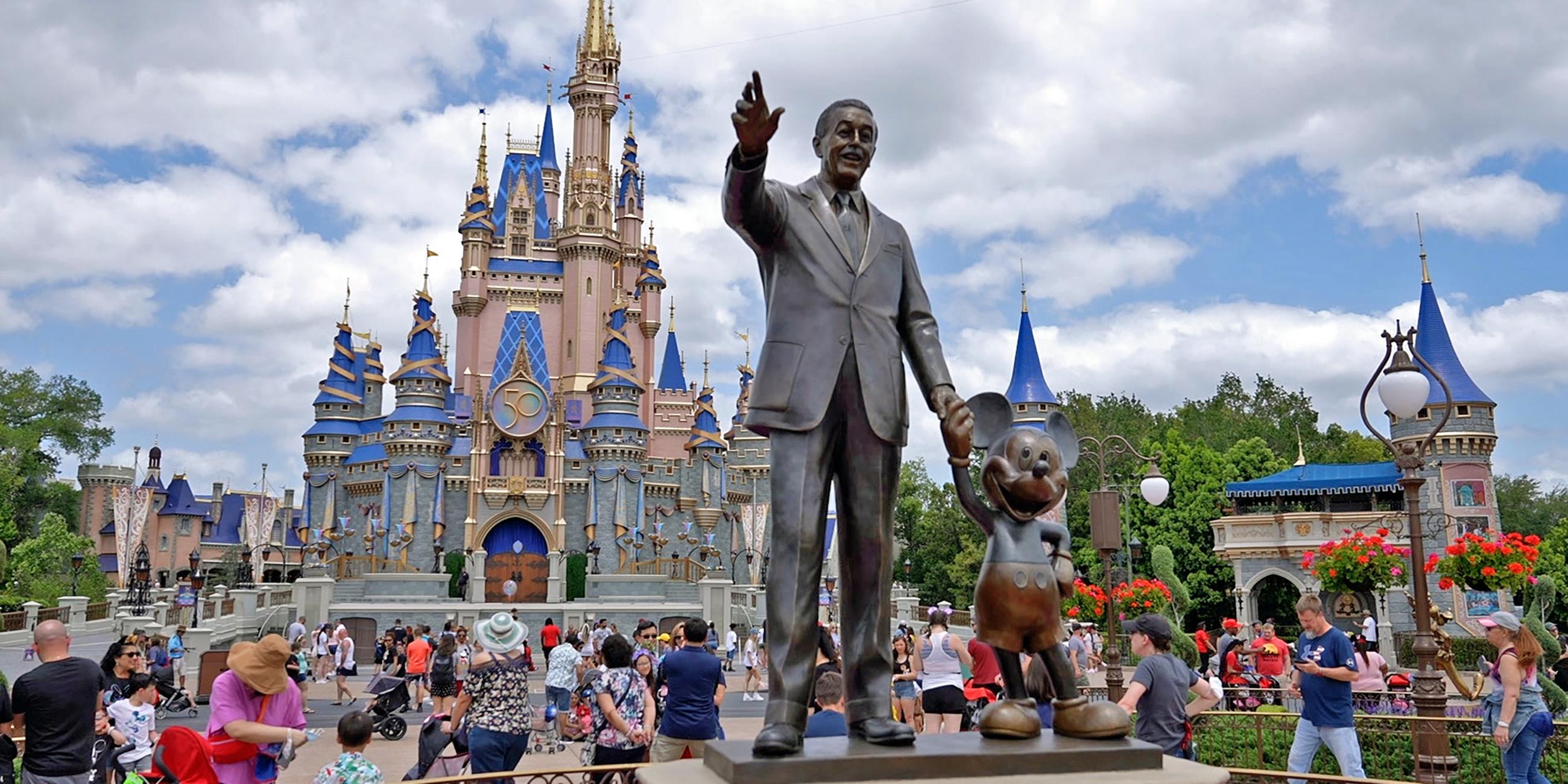 Eine Statue von Walt Disney, der Micky Maus an der Hand hält, steht in einem Vergnügungspark. Dahinter laufen dutzende Touristen vor einem Märchenschloss.