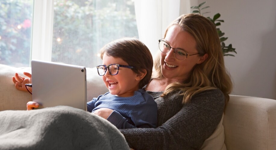 Adult and child wearing glasses, smiling while looking at a tablet on a couch.