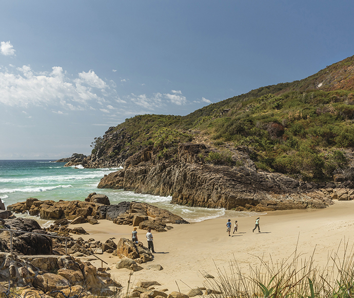 Scenic view of Little Bay, located in Arakoon National Park. The water gentle rolls onto the light coloured sand, with a series of adults and children walking across the beach. Grassy hills and trees line the background, while rocks of varying sizes are scattered across the shore.