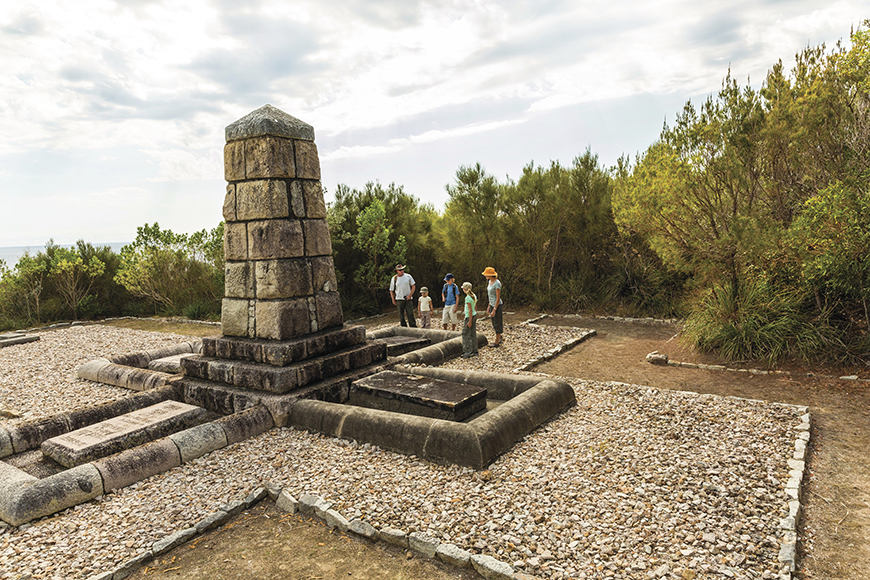 A World War 1 monument as part of the Monument track sits in the centre, with a family of visitors exploring the monument. The monument has been precisely crafter out of a variety of stones, creating a striking geometric shape, while trees line the perimeter of the monument.