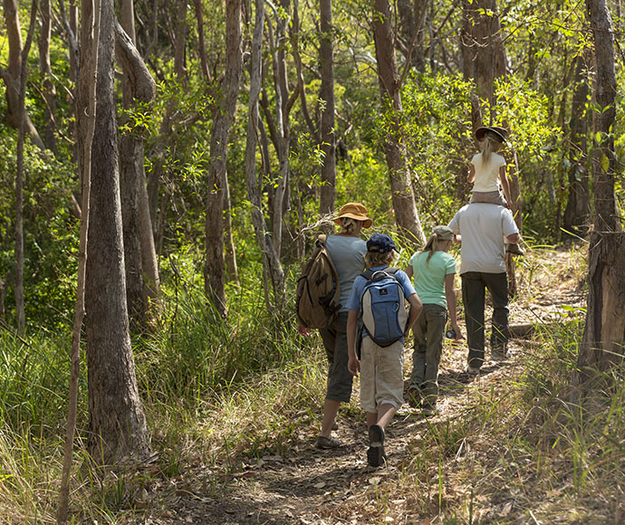 A young family walks across the Powder Magazine Trail in Arakoon National Park. There are three children, with one child on the shoulders of a male figure, while the remaining two children walk close to the female figure in the photo. Sunlight shines through the tall trees and illuminates the vibrant light green of the leaves.