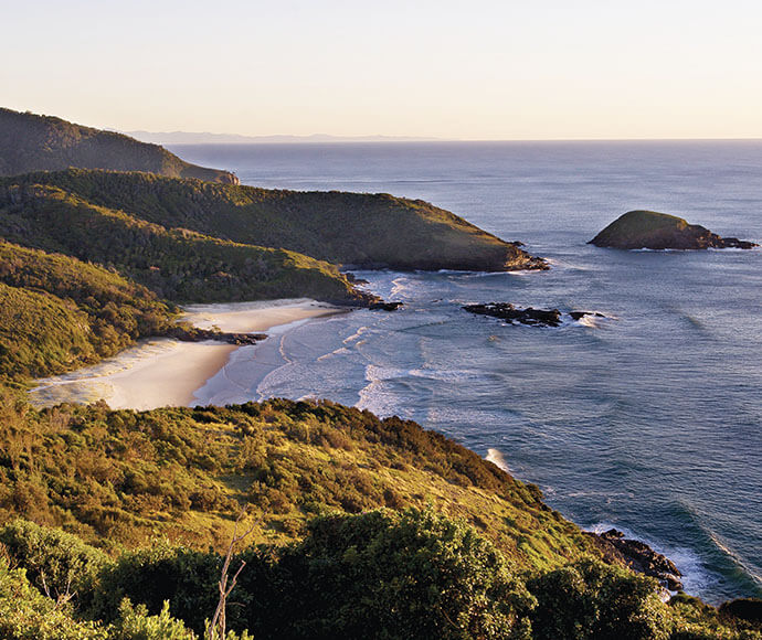 A scenic coastal landscape featuring a beach with white sand, surrounded by lush green hills and cliffs. The ocean waves gently lap against the shore, with rocky outcrops extending into the water. An island is visible in the distance, adding depth to the scene. The image is taken from an elevated viewpoint, providing a broad perspective of the coastline and surrounding natural beauty. The lighting suggests it is either early morning or late afternoon, casting a warm glow over the landscape.