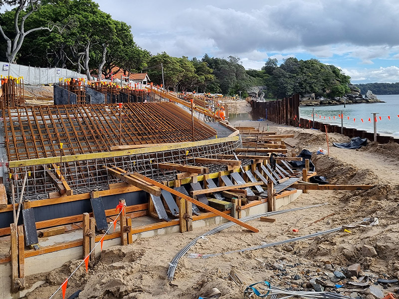 A construction site featuring a large curved structure supported by wooden beams, set against a backdrop of trees and an overcast sky