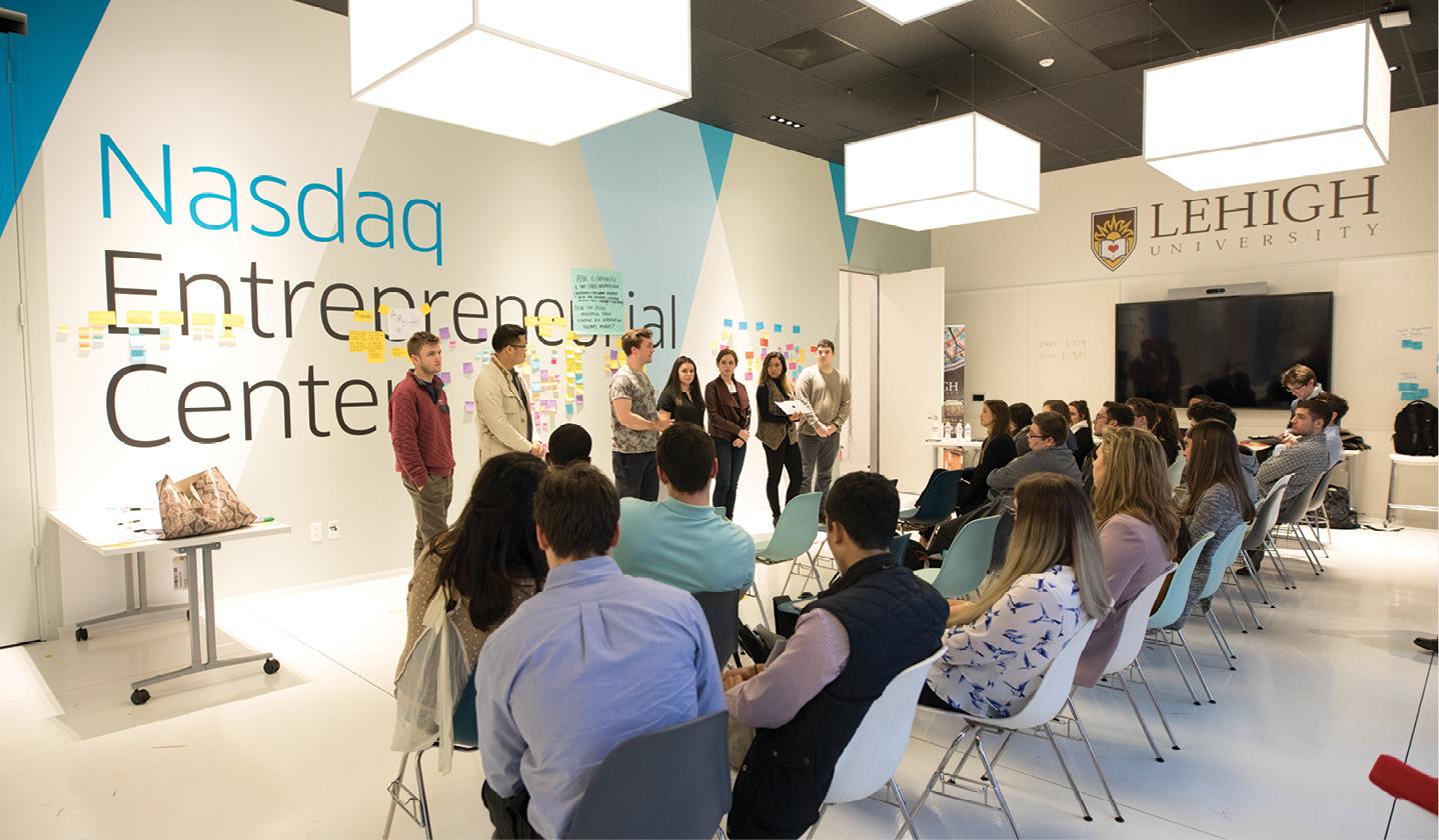 Several people are seated and listening to a presentation in a modern conference room. A group of speakers stands near a large screen and next to a wall displaying "Nasdaq Entrepreneurs Center." Attendees are facing towards the speakers.